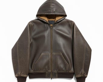 May include: A brown leather hooded jacket with a full zipper closure and a front pocket. The jacket has a brown ribbed hem and cuffs. The interior of the hood is lined with a tan, fuzzy material. The jacket is displayed on a white background.