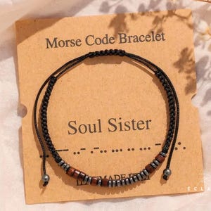 Op de afbeelding: Een zwart Morsecode-armband met houten en zilveren kralen, gepresenteerd op een bruine kaart. De kaart leest "Morse Code Bracelet" en "Soul Sister". De armband is verstelbaar en heeft een delicaat ontwerp.