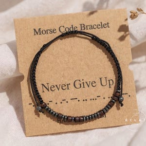 Könnte beinhalten: Ein schwarzes Morsecode-Armband mit Holz- und Metallperlen, präsentiert auf einer braunen Karte. Die Karte trägt die Aufschrift "Morse Code Bracelet" und "Never Give Up". Das Armband befindet sich auf neutralem Hintergrund.