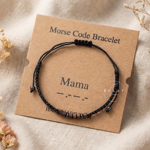 Op de afbeelding: Een zwart Morsecode-armband met het woord "Mama" in kralen. De armband wordt gepresenteerd op een bruine kaart met de tekst "Morse Code Bracelet". De kaart ligt op een beige oppervlak.