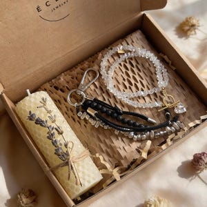 May include: A gift box containing a honeycomb candle with lavender, a clear crystal necklace, and a black and silver keychain. The box is made of cardboard and has the text "ÉCLAT JEWELRY" printed on the lid.