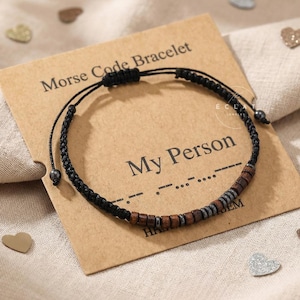 Op de afbeelding: Een zwarte Morsecode-armband met houten en grijze kralen wordt getoond op een bruine kaart met de tekst "Morse Code Bracelet" en "My Person". De armband ligt op een neutraal gekleurd oppervlak met hartvormige confetti.