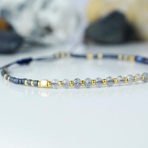 May include: A delicate bracelet featuring a combination of dark gray, gold, and clear beads. The bracelet includes small gold accents and a section of blue beads. The adjustable cord is dark blue.
