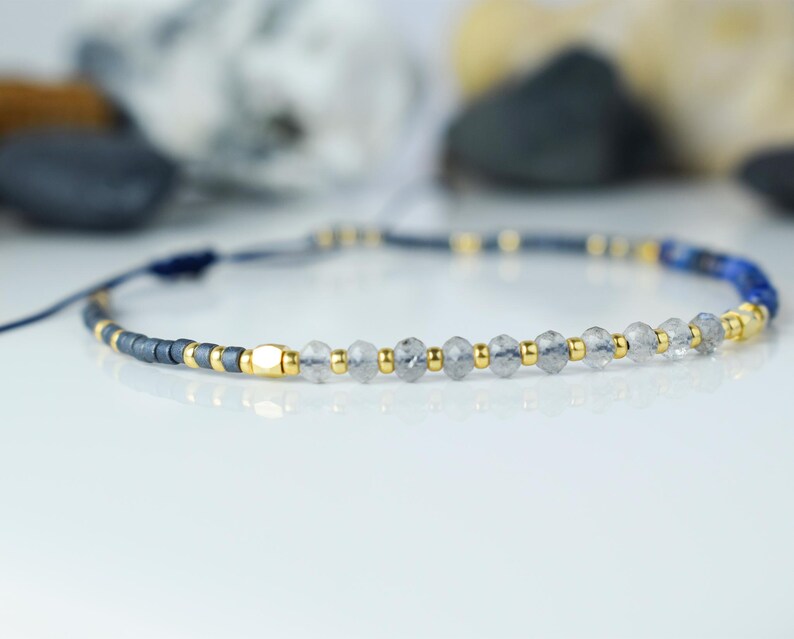 May include: A delicate bracelet featuring a combination of dark gray, gold, and clear beads. The bracelet includes small gold accents and a section of blue beads. The adjustable cord is dark blue.