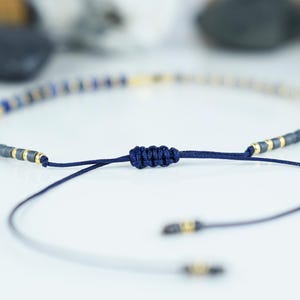 May include: Adjustable bracelet featuring a string of small, dark blue and gold-colored beads. The bracelet is tied with a dark blue cord, and the ends have small gold accents. The bracelet is designed to be adjustable.