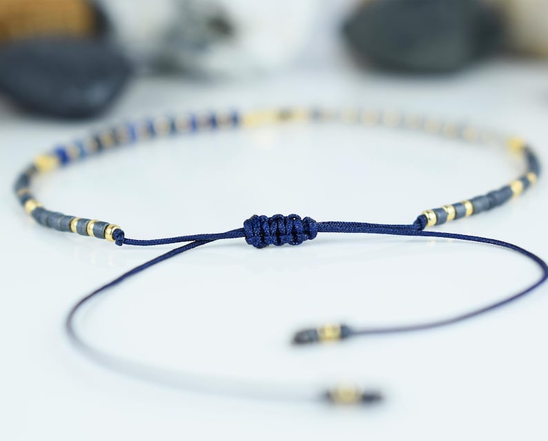 May include: Adjustable bracelet featuring a string of small, dark blue and gold-colored beads. The bracelet is tied with a dark blue cord, and the ends have small gold accents. The bracelet is designed to be adjustable.