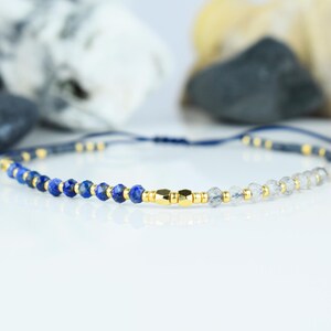 May include: A delicate bracelet featuring a combination of blue and clear gemstone beads, interspersed with small gold-colored beads. The bracelet is strung on a dark blue cord and has a gold-colored clasp. The jewelry is displayed on a white surface.