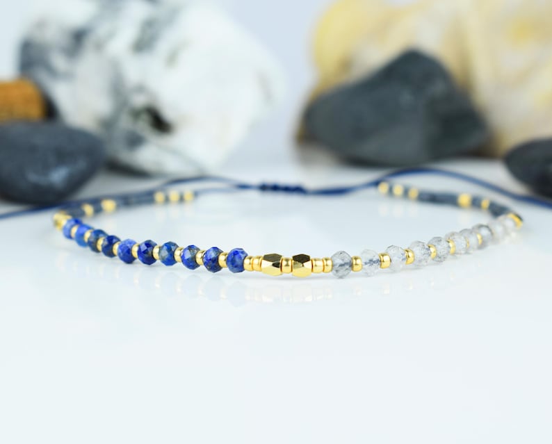 May include: A delicate bracelet featuring a combination of blue and clear gemstone beads, interspersed with small gold-colored beads. The bracelet is strung on a dark blue cord and has a gold-colored clasp. The jewelry is displayed on a white surface.