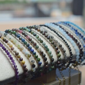 May include: A display of beaded bracelets in various colors, including purple, green, blue, and black. The bracelets are made of small, round beads and are arranged on a white, cushioned surface.