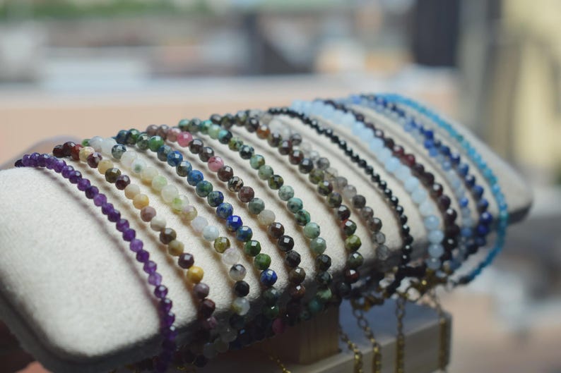 May include: A display of beaded bracelets in various colors, including purple, green, blue, and black. The bracelets are made of small, round beads and are arranged on a white, cushioned surface.