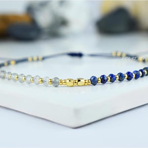 May include: A delicate bracelet featuring a combination of clear and blue gemstone beads, interspersed with gold-colored metallic accents. The bracelet is strung on a dark blue cord, with the brand name "ECLAT JEWELRY" visible.
