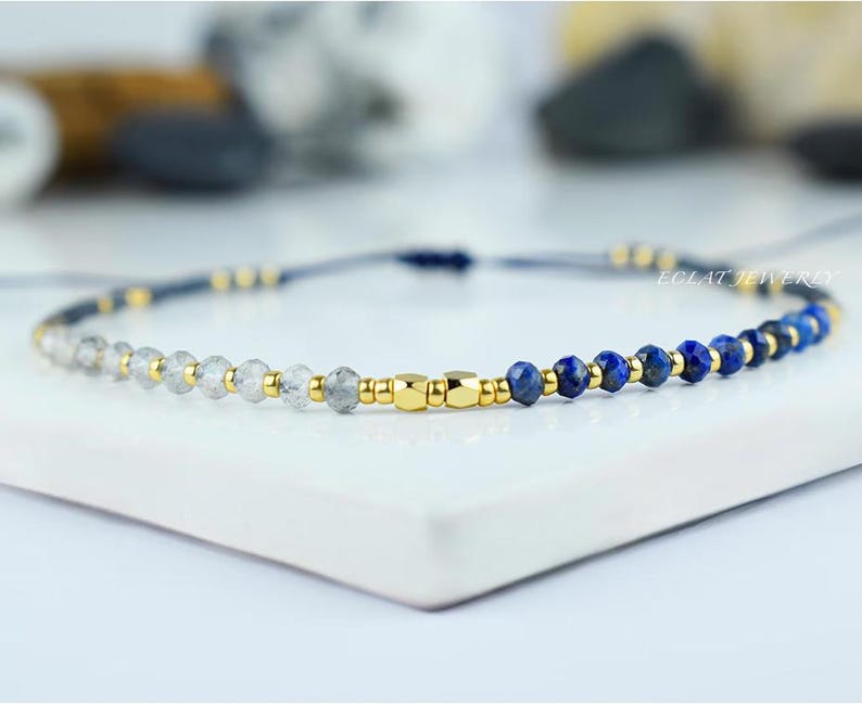May include: A delicate bracelet featuring a combination of clear and blue gemstone beads, interspersed with gold-colored metallic accents. The bracelet is strung on a dark blue cord, with the brand name "ECLAT JEWELRY" visible.