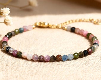 May include: A delicate bracelet featuring a strand of multi-colored gemstone beads. The beads are in shades of pink, green, blue, and brown, strung together with a gold-colored chain and clasp. The jewelry is displayed on a neutral-toned surface.
