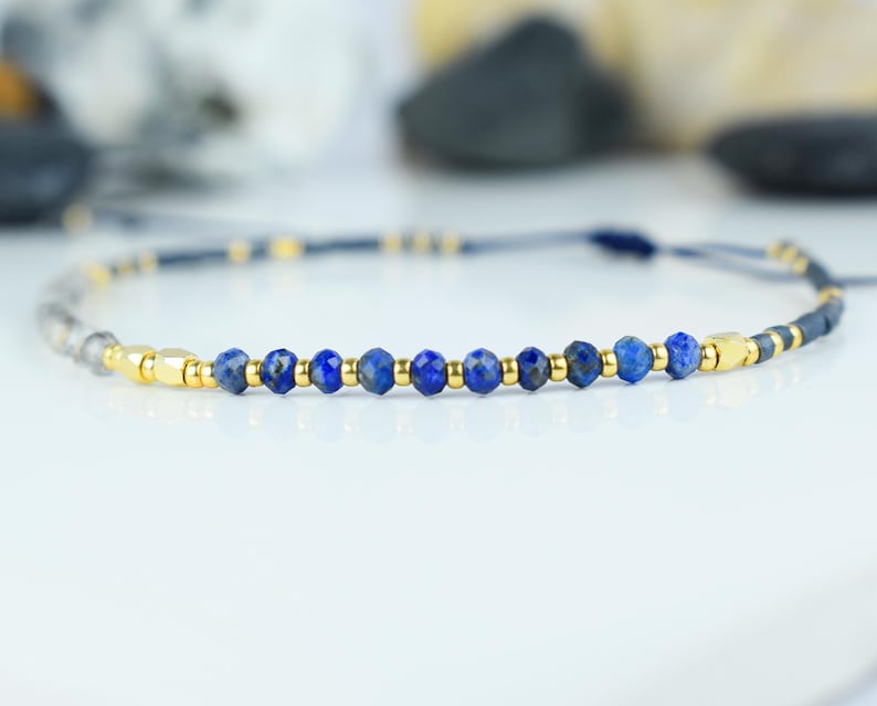 May include: A delicate bracelet featuring faceted blue lapis lazuli beads interspersed with gold-colored metal accents. The adjustable cord is a dark blue-gray color, creating a minimalist and elegant design. The bracelet is suitable for everyday wear.