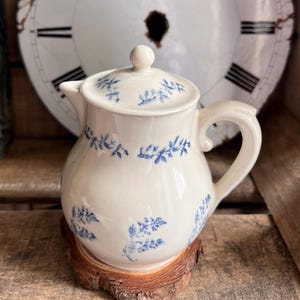 以下が含まれることがあります： 繊細な青い花柄が施されたクリーム色の陶器製クリーマーまたは小さなピッチャー。丸みを帯びた本体、曲線的なハンドル、小さなつまみ付きの蓋が特徴です。天然木の輪切りの台座の上に置かれています。