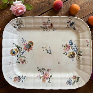 Può includere: Un vassoio da portata rettangolare in ceramica color bianco sporco con bordi smerlati, caratterizzato da un delicato motivo floreale con illustrazioni botaniche rosa, blu, gialle e verdi sparse al centro e lungo il bordo scanalato.
