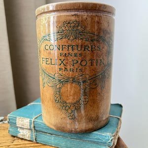 May include: A cylindrical brown stoneware jar with a glossy finish and a dark green printed label that reads CONFITURES FINES FELIX POTIN PARIS. The jar features an ornate decorative border surrounding the text and a circular emblem below.