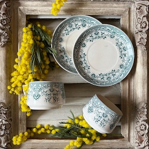 Puede incluir: Un juego de té vintage con un diseño floral en verde azulado y blanco, que incluye dos platillos y dos tazas. El juego está dispuesto con flores amarillas de mimosa dentro de un marco de madera decorativo, ornamentado y envejecido.