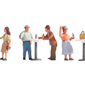 May include: A set of miniature plastic figurines depicting six people and a small dog in a social setting. The figures include individuals in casual clothing standing around three small round tables, with one person reaching under a table.