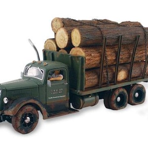 May include: A green vintage-style toy logging lorry with a weathered finish, carrying a load of brown cylindrical logs secured by chains. A small figurine wearing a red shirt and blue trousers stands beside the lorry, which features a driver inside.
