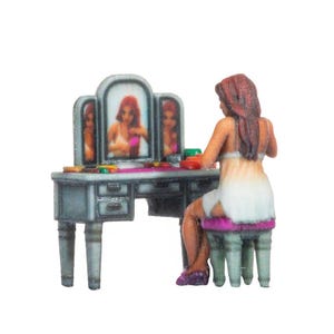 May include: A miniature figurine of a person with long brown hair sitting on a purple stool at a grey vanity table with a tri-fold mirror. The person wears a white garment and is reflected in the mirror, which shows small colorful items on the table.