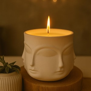 May include: A white candle in a ceramic holder shaped like a face. The candle is lit, with a flame burning brightly. The holder sits on a wooden base, with a small potted plant to the side. The background is a warm, neutral color.