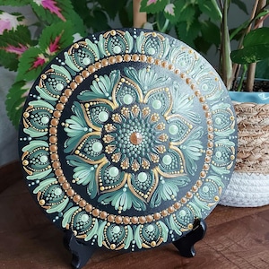 May include: A round, black decorative plate with a hand-painted mandala design. The mandala features a central floral pattern in shades of green and gold, surrounded by intricate dot patterns. The plate is displayed on a black stand.