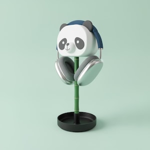 Op de afbeelding: Een witte koptelefoonstandaard in de vorm van een panda, met een blauwe hoofdband en grijze oren. De standaard houdt zilveren koptelefoons vast en wordt ondersteund door een groene bamboestengel en een zwarte cirkelvormige basis. De achtergrond is lichtgroen.