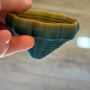 Puede incluir: Objeto impreso en 3D de color verde azulado y dorado, con forma cónica. El objeto tiene una serie de crestas horizontales y un borde superior festoneado. El degradado de color cambia de verde azulado a dorado, creando un efecto visual interesante.