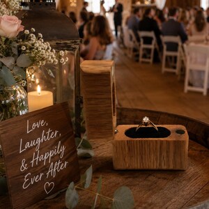 May include: A gold engagement ring with a single diamond sits in an open wooden box on a barrel. Next to it is a dark wooden sign with the white text "Love, Laughter, & Happily Ever After" and a heart, a glass jar with flowers, and a black lantern.