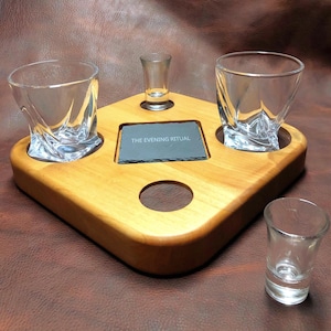 May include: A wooden serving tray with two square-shaped glasses, two shot glasses, and a small slate tile that reads "THE EVENING RITUAL". The tray is made of light-colored wood and has a diamond shape.