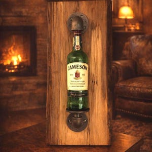 May include: A rustic wooden wall-mounted whiskey bottle holder. The holder features a green Jameson bottle secured by industrial pipe fittings. The wood has a natural grain, and the background includes a fireplace and warm lighting.