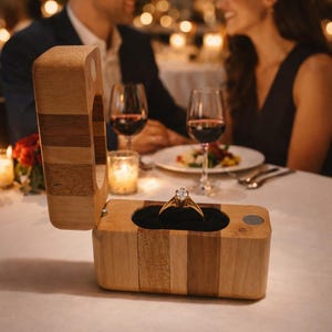 May include: A wooden ring box with a gold engagement ring inside, open on a white tablecloth. The box is made of light and dark wood panels. A couple is seated at a table in the background, with wine glasses and candles.