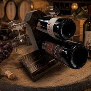 May include: A dark stained wooden wine rack holding two wine bottles and two wine glasses. The rack is angled, with the bottles and glasses displayed at an angle. The background includes wine barrels and a bottle of wine.