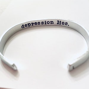 May include: A silver-colored cuff bracelet with the stamped text "depression lies." The open-ended bracelet is made of a smooth, shiny metal. The text is in a simple, sans-serif font.