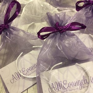 May include: Multiple purple and white organza bags with silver bracelets inside. The bags are tied with purple ribbon and the bracelets have the text "Ann Evangeli Hand Stamped Jewelry" on them.