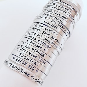 May include: A stack of silver bracelets with inspirational messages engraved on them. The messages include "I am Loved", "follow your arrow", "You are my Sunshine", "It Takes a Village", "blessed", "I am STRONG", "follow your bliss", "FIGHTER", "DREAM BIG", "enlighten & educate", "fearless", "for the Stars", "Connecticut", "FOOTBALL MOM", "I am Loved", "your dream", "enlighten & educate".