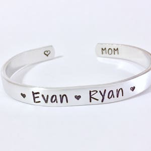 May include: Silver bangle bracelet with the words "MOM", "Evan", and "Ryan" engraved on it, separated by heart symbols.
