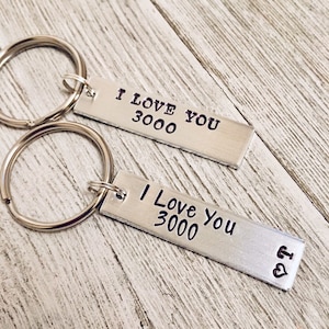 May include: Two silver keychains with the phrase "I Love You 3000" engraved on them. The keychains are on a wooden surface.
