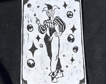 May include: Black and white linocut print of a jester figure with a pointed hat and a jester's costume. The jester is surrounded by small circles and stars.