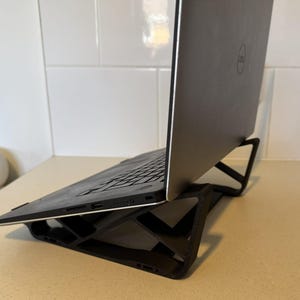 May include: A black laptop computer is positioned on a black, geometric laptop stand. The stand is designed to provide an angled surface for the laptop, improving ergonomics. The laptop is a Dell model.