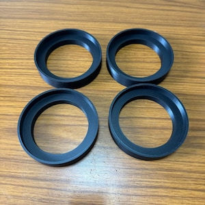 Puede incluir: Cuatro anillos negros, circulares, impresos en 3D están dispuestos sobre una superficie de madera. Cada anillo tiene un borde biselado y un centro hueco. Los anillos son idénticos en tamaño y forma y parecen ser de plástico.