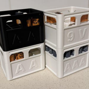 May include: Two battery storage containers, one black and one white, stacked together. The white container has "AAA" and "9 V" labels. The black container is stacked on top of the white container. Batteries are visible inside the containers.