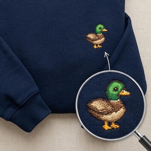 May include: A navy blue crewneck sweatshirt featuring a small embroidered mallard duck on the chest. The duck has a green head, brown body, and yellow feet. A circular magnifying glass overlay shows a detailed view of the embroidery stitches.