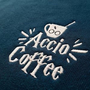 May include: A dark teal sweatshirt featuring white embroidery. The text reads Accio Coffee in a stylized font. Above the text is an embroidered coffee mug with a lightning bolt and round glasses design, accented by small radiating lines.