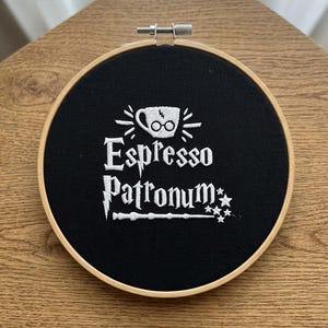 May include: A circular wooden embroidery hoop containing black fabric with white embroidery. The design features a coffee cup with glasses and a lightning bolt, the text "Espresso Patronum," a lightning bolt, a magic wand, and three small stars.