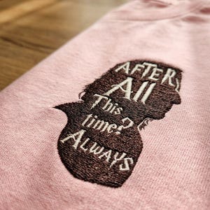 May include: A light pink sweatshirt featuring a dark brown embroidered silhouette of a head in profile. The embroidery includes the text "After All This time? ALWAYS" in a white serif font, with a lightning bolt symbol integrated into the letter A.