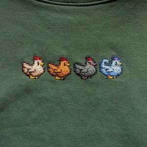 May include: A dark green sweatshirt featuring four embroidered chickens in a row. The chickens are in shades of white, orange, gray, and blue, each wearing a red cap. The sweatshirt has a crew neck.