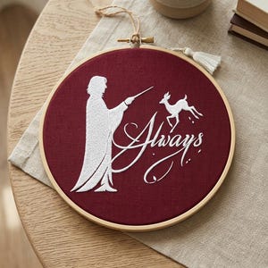 May include: A circular wooden embroidery hoop containing deep red fabric with white thread embroidery. The design features a silhouette of a person holding a wand, a leaping deer, and the word Always in a cursive script, finished with a small tassel.