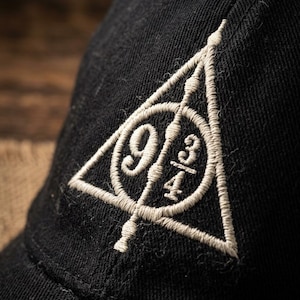 May include: A black baseball cap featuring an embroidered design in light beige thread. The embroidery depicts a triangle enclosing a circle and a vertical line, with the numbers 9 and 3/4 positioned inside the circle, forming a graphic symbol.